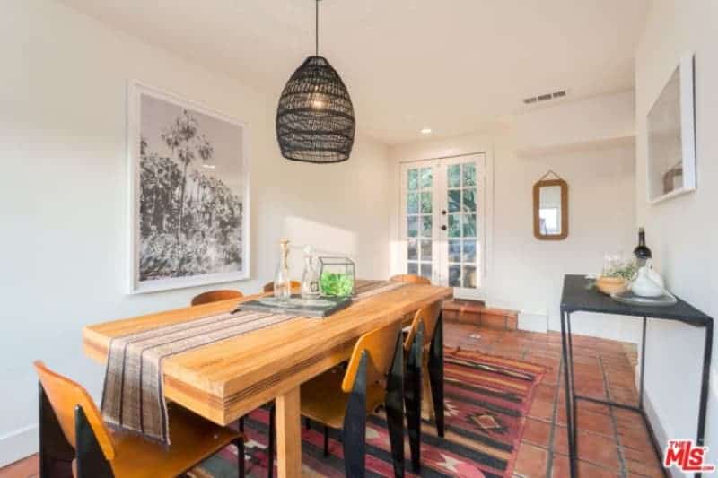 The terracotta flooring of this dining room provides an earthy hue to contrast the white walls and ceiling with a pendant light that has a black woven basket for a hood. This hangs over the thick wooden dining table that is paired with wooden chairs and accented with a colorful area rug underneath as a complement to the flooring.