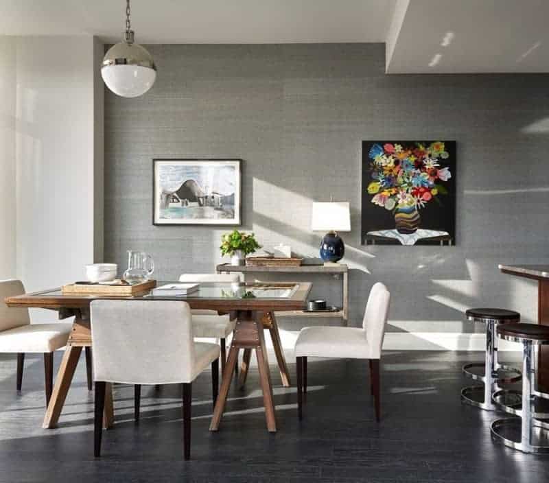 A modern spherical pendant light hangs over the wooden dining table with a glass panel on top. This is paired with dining chairs with dark wooden legs and white cushions that stand out against the gray wall adorned with a console table and a couple of colorful paintings.