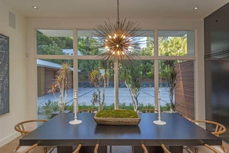 The black rectangular wooden dining table stands out against the light wooden wishbone chairs and the hardwood flooring that are illuminated by the yellow lights of the peculiar pendant light with a sun-like design. This yellow light is augmented by the natural lights coming in from the glass wall.