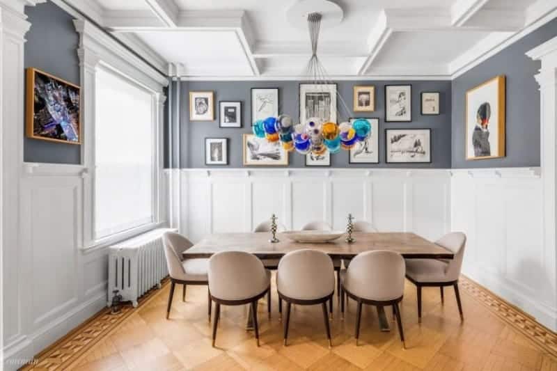 The bright wainscoting of this medium-sized dining room matches with the white coffered ceiling and is complemented by the hardwood flooring that has a subtle checkered design in the middle bordered with intricate designs. This goes well with the beige cushions of the dining chairs paired with the wooden table topped with colorful pendant lights.