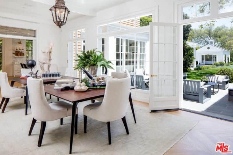 The patio scene outside the French glass windows and glass doors bring in natural lighting that brightens up the white ceiling with exposed beams that support a classic lantern-like  pendant light that that brings a certain Spanish-style flair to the bright aesthetic.