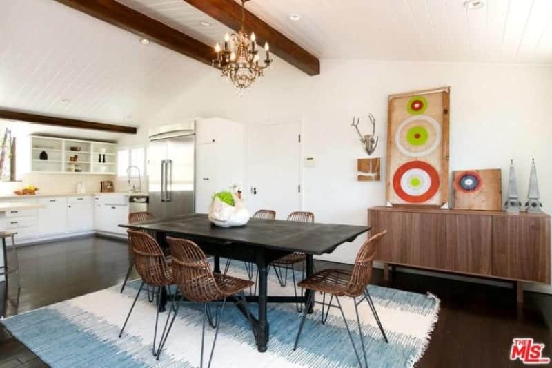 The bright and white ceiling has a couple of wooden exposed beams that matches with the hue of the rustic chairs surrounding the black wooden dining table that stands out against the light hue of the area rug on the dark hardwood flooring.