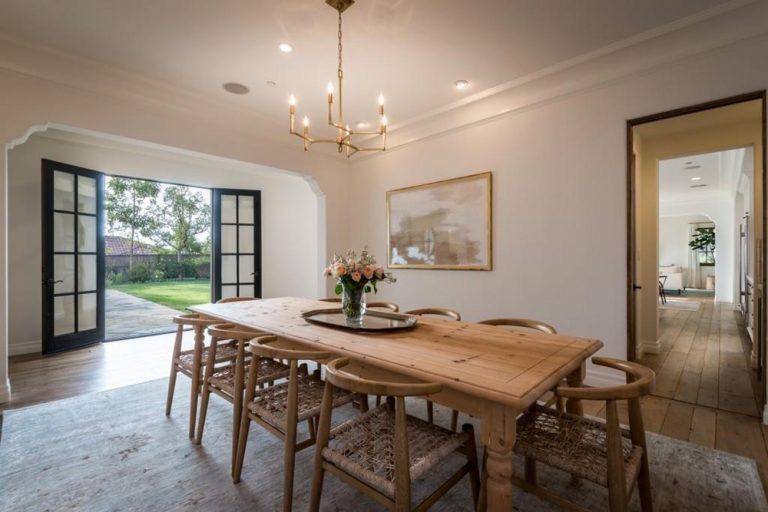 The rustic wooden chairs with woven wicker seats fit perfectly with the wooden dining table topped with a small lovely golden chandelier that casts warm yellow light on the beige ceiling and walls adorned with a colorful painting.