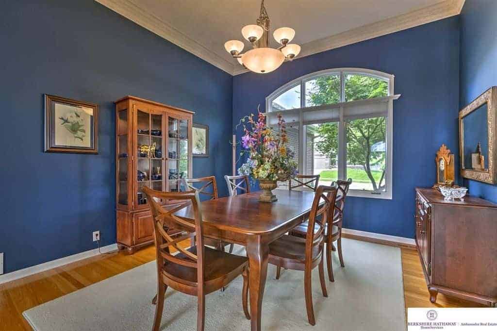 The charming blue walls are brightened and enhanced by the large arched window that brings in natural lighting that augments the warm chandelier over the wooden dining table that matches with the cabinet and hardwood flooring topped with a light gray area rug.