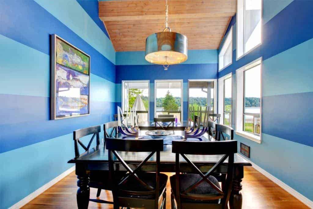 The delightful blue-striped walls offers a bright and cheerful contrast to the traditional dark wooden table and its wooden cross back chairs that complement the hardwood flooring. These are all brightened by the natural lights coming from the windows.