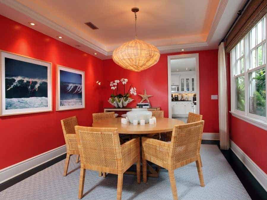 This beach-style dining room has bright red walls adorned with a couple of framed photos depicting surfers riding a large wave. Adjacent to it is a console table with flowers planted on a large seashell beside a woven wicker starfish that matches the dining set and the pendant light.