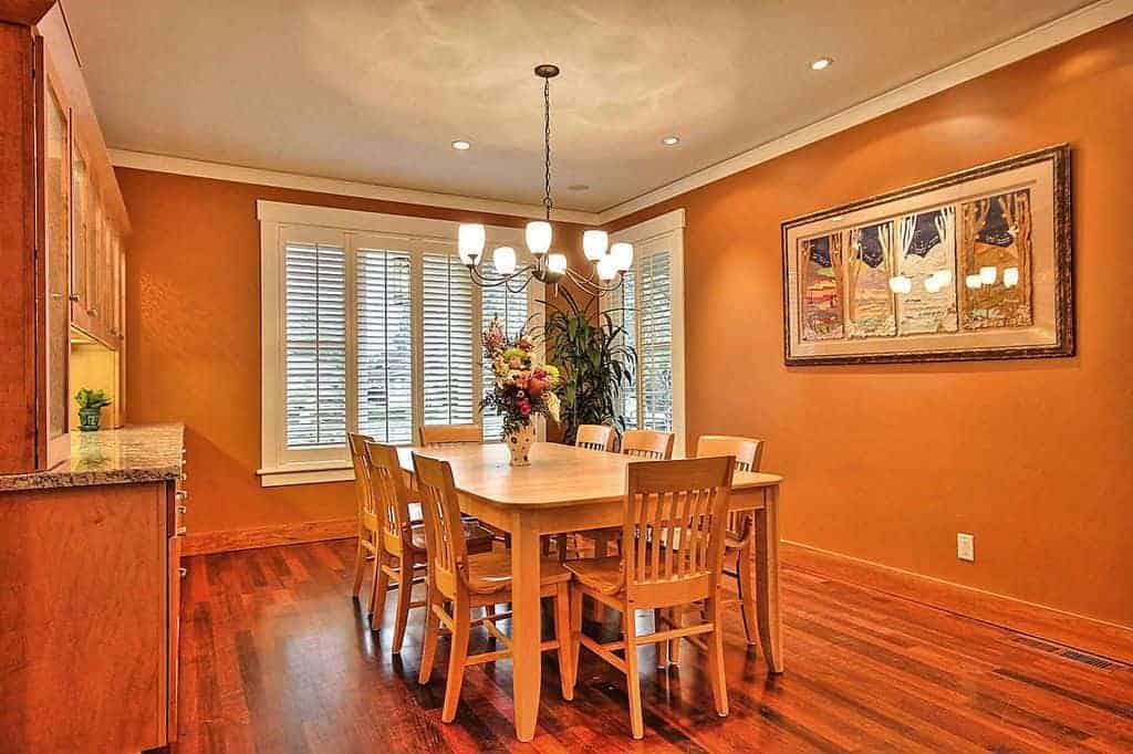 The orange walls seem to blend with the rest of the dining room from the hardwood flooring to the simple wooden dining set in the middle of the room under a simple chandelier. This is brightened by the white shuttered windows and the ceiling of this medium-sized dining room.