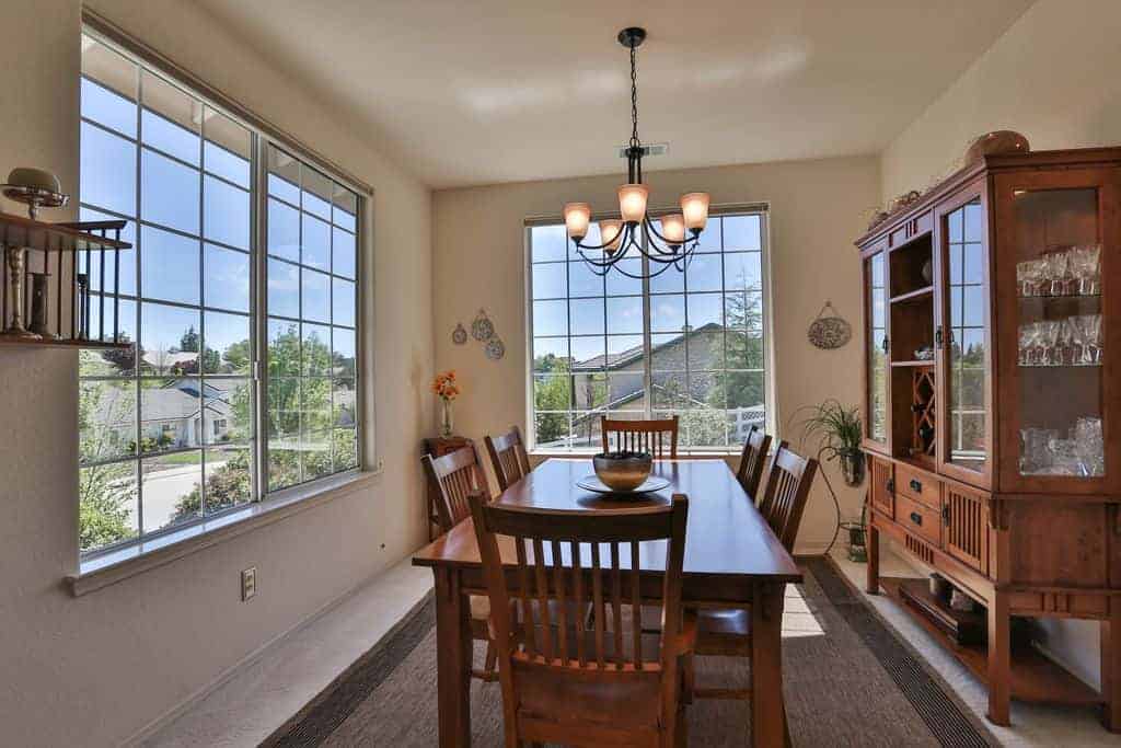 The large French windows of this dining room illuminates the earthy wooden hues of the dining table and slat back chairs that matches with the dining room cabinet that has glass panels on it showcasing the crystal ware and wines against a light yellow wall.