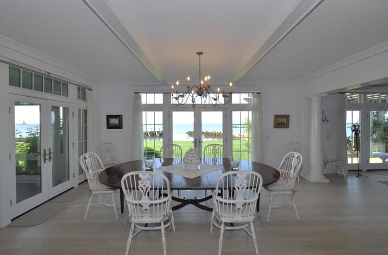 The classic white wooden chairs that stand out against the dark wooden elliptical dining table matches with the light hardwood flooring, white walls and white ceiling that has exposed wooden beams flanking a thin chandelier.