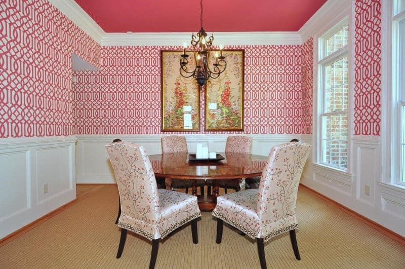 The matte pink ceiling that supports a wrought iron dark chandelier matches with the patterned wallpaper of the upper walls that provide a nice complex background for the white wainscoting, wooden round dining table and the cushioned chairs with floral slip covers.