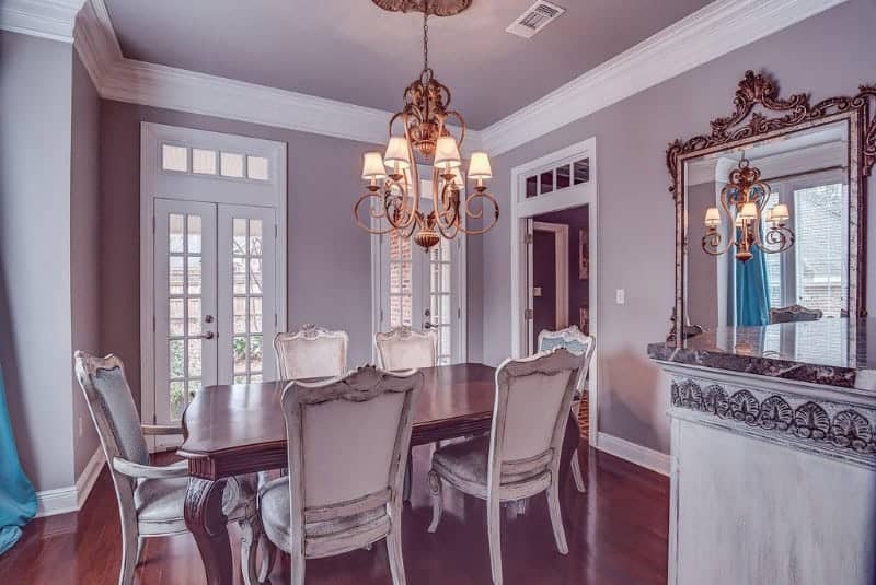 This dining room has a classic quality to its antique wooden dining chairs that stand out against the redwood dining table that blends with the hardwood flooring. This is augmented by the warm chandelier that stands out against the purple-gray walls.