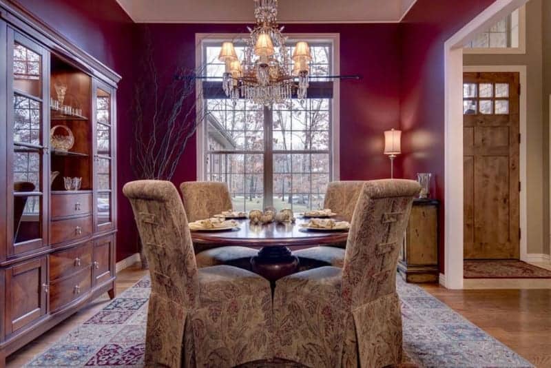 The dark reddish purple tone of the walls are augmented by the warm yellow lights of the table lamp and the matching chandelier. This is also brightened by the natural lights coming in from the tall windows onto the floral patterns of the slipcovers and area rug.