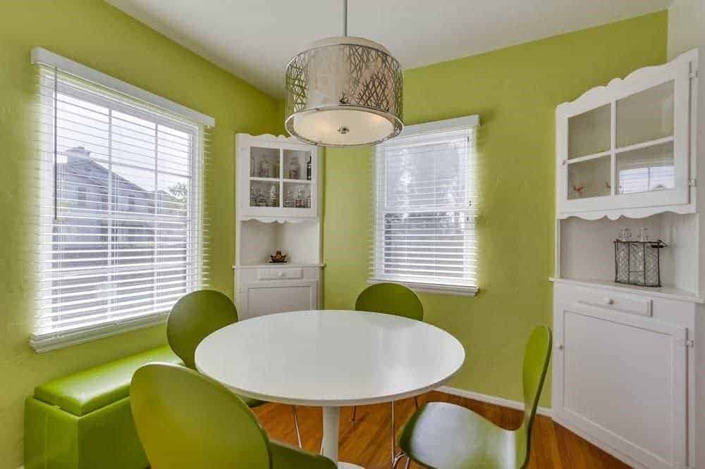 The light green walls that are brightened by the couple of windows with white blinds are a complement to the the dark green modern chairs that surround the white modern round dining table. It also matches the green leather cushioned bench by the window.