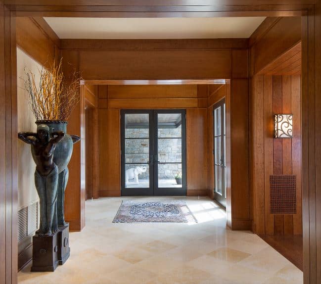 The gray frame of the wooden door supports glass panels that bring in an abundance of natural lighting to the wooden walls and matching wooden ceiling complemented by the beige marble flooring that is contrasted by dark statuettes.