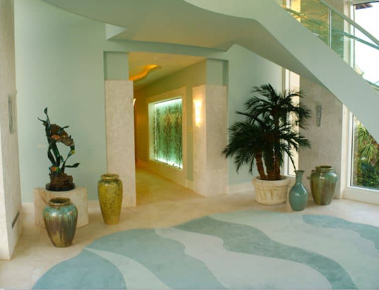 Layout: I simply adore this open and airy foyer layout that features nothing but tropical plants and gorgeous pottery. The room is dedicated to simply guiding you to the rest of what is obviously an incredible tropical home. Furniture: This foyer features no furniture, and instead acts almost as a view room where you will swiftly be whisked into another, even more incredible room. Flooring: The floor in this room is truly something to behold, featuring incredibly painted stone tiling that is meant to evoke the feeling of wading in shallow water. This is so unique, so lush, and simply incredible to look at. Lighting: While there isn't much featured lighting in the foyer itself, the lighting in the hallway feels very exciting. The warm hues of the lights really entice you to walk through the gorgeous under-waterway.Decor: Tropical plants adorn the room, while a very smart paint job further emphasizes the feeling that you are calmly floating in shallow, warm waters. The perfect oasis that is this foyer was designed by Dezign Inspirations/ Home Design Resource.