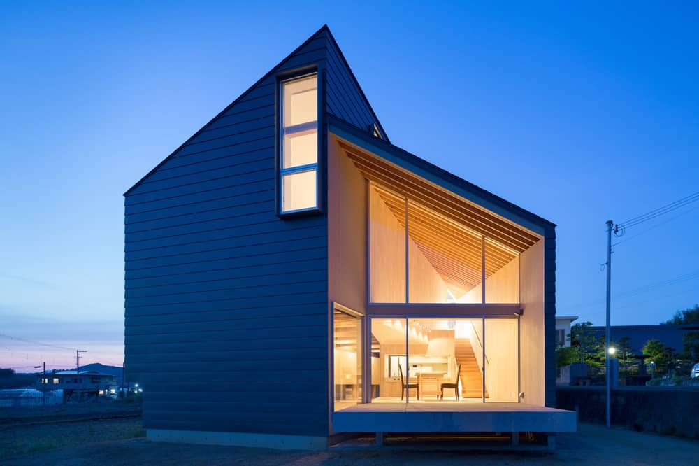 This is a nighttime view of the back of the house featuring an asymmetric design paired with a large glass wall that shows the interiors of the kitchen.