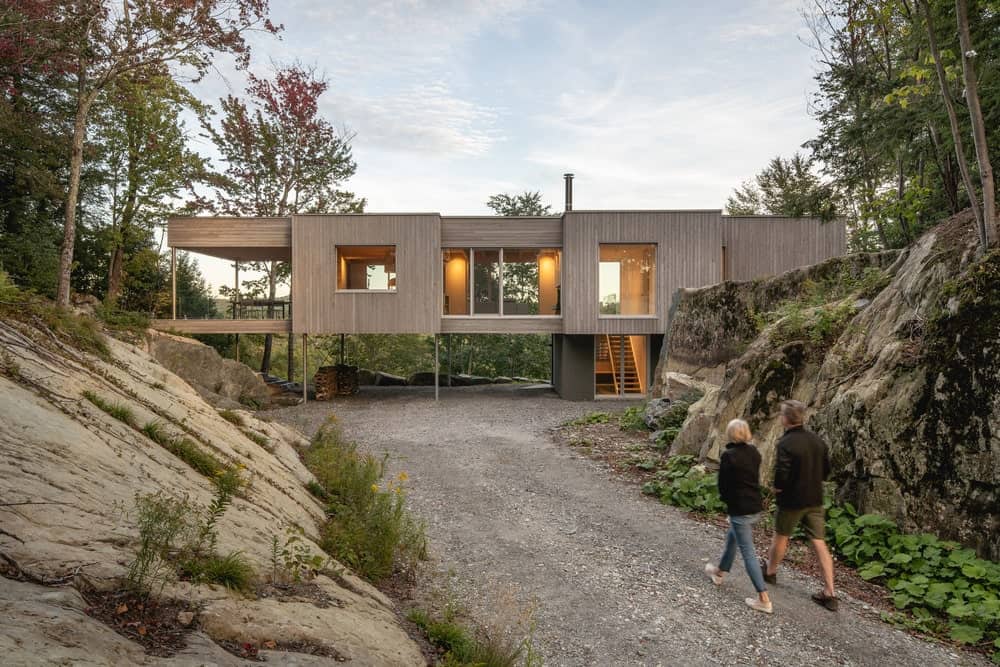 This is a view of the house from the driveway that leads to the wooden structure with glass windows and a large covered patio on the far left side.