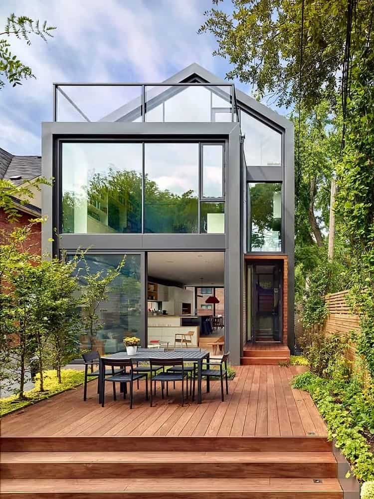 This is a look at the back of the house that is dominated by glass walls. These are complemented by the wooden deck floor of the backyard flanked by lush landscaping and topped with a gray outdoor dining set.