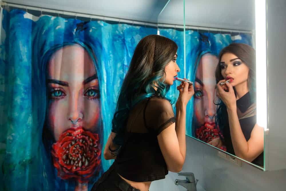 A woman doin her make-up in a bathroom that has an artistic shower curtain.