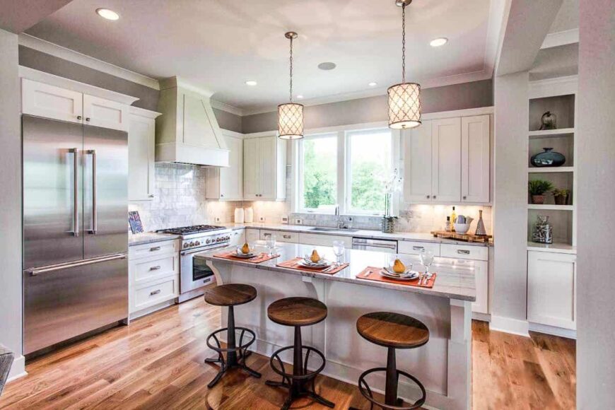 The kitchen is equipped with stainless steel appliances, white cabinetry, and a breakfast island complemented by round bar stools and a pair of glass pendants.