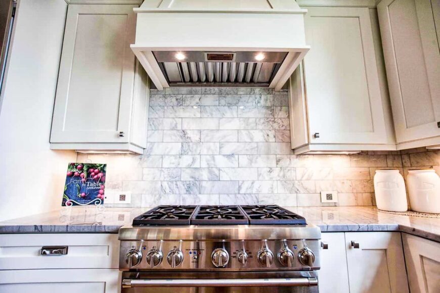 A closer look at the stainless steel cooking range under the bespoke vent hood that's fixed against the marble subway tile backsplash.