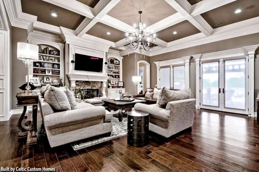 The living room has polished hardwood flooring, a pair of french doors, and a coffered ceiling mounted with a lovely chandelier and recessed lights.