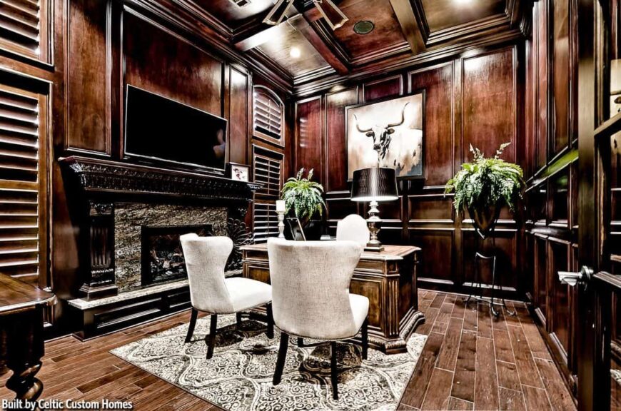 Home office with a warm fireplace, wingback chairs, a wooden desk, and dark wood coffered ceiling matching with the wood-paneled walls adorned by a bull artwork.