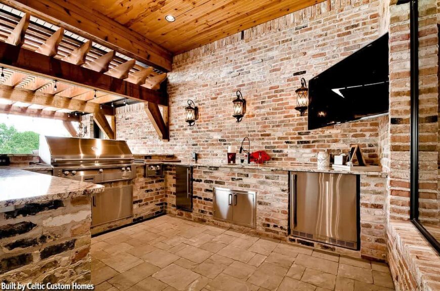 Outdoor kitchen with stainless steel appliances and cabinets along with a corner TV fixed against the brick walls.