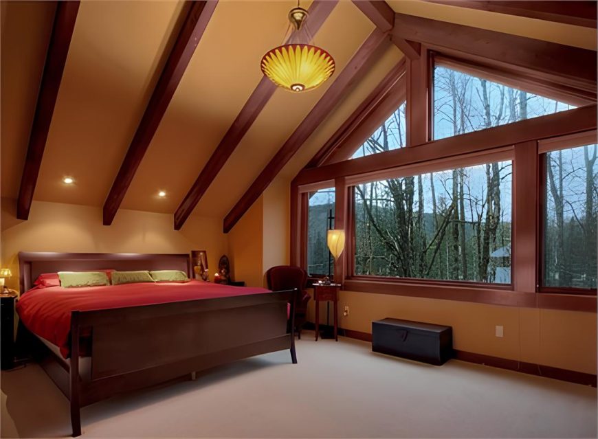 The primary bedroom has beige carpet flooring, wooden framed windows, and a cathedral ceiling lined with rustic beams.