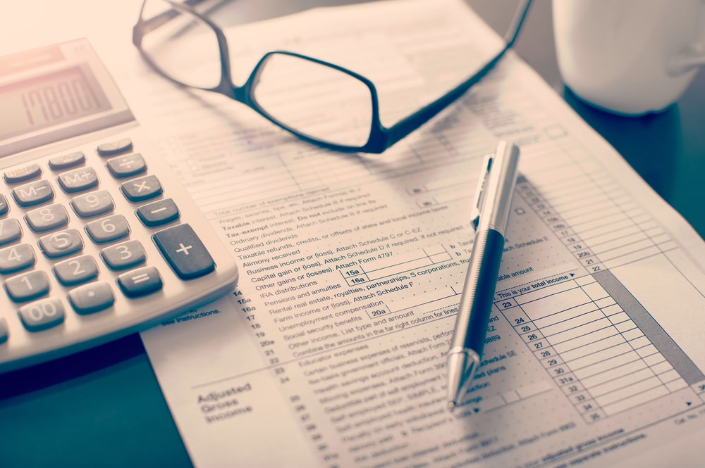 Income tax return form beside a calculator, eyeglasses, a pen, and a mug.