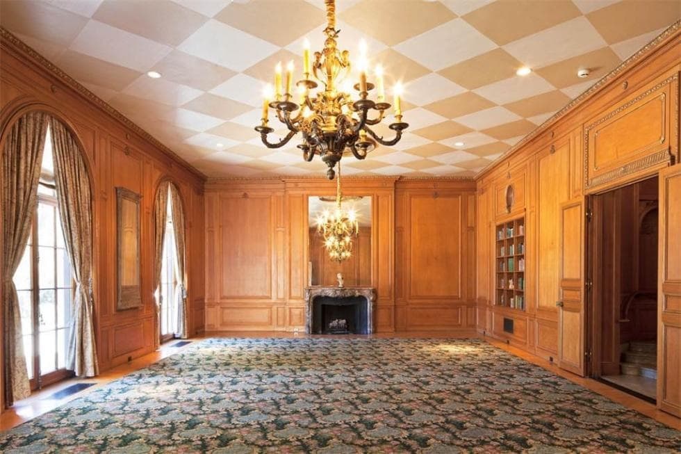 This is the spacious wood-paneled library with a checkered ceiling that hangs a chandelier in the middle of the spacious room that has a fireplace on the far side. Image courtesy of Toptenrealestatedeals.com.