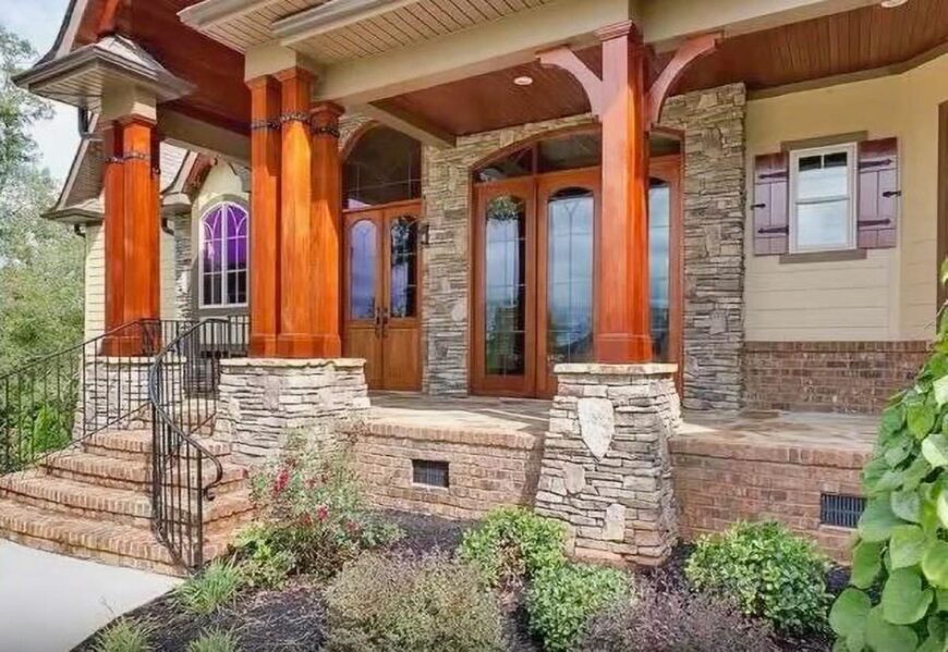 Covered front porch showing the wooden columns, glazed entry door, stone brick accents, and a stoop with iron railings.