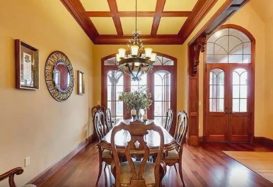 Formal dining room with a wooden dining set, hardwood flooring, and warm yellow walls adorned by an intricate round mirror and framed artworks.