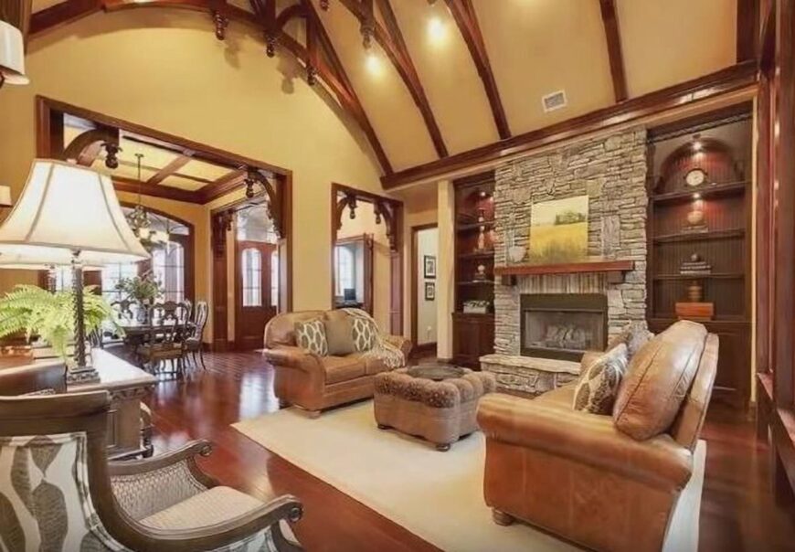 Living room with high beamed ceiling, leather couches, tufted ottoman, and a stone fireplace flanked by wooden built-ins.Living room with high beamed ceiling, leather couches, tufted ottoman, and a stone fireplace flanked by wooden built-ins.