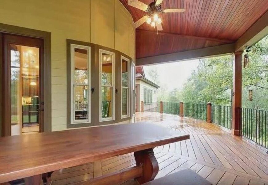 Covered porch with wide plank flooring, wrought iron railings, wooden table, and a ceiling fan mounted on the wood-paneled ceiling.