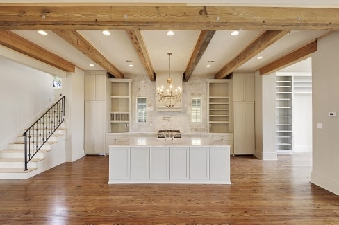 The kitchen has a beamed ceiling, white cabinets, and a marble top island lit by a candle chandelier.