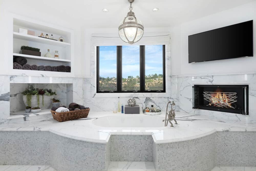 This is the bright white bathroom with a bathtub under the window. On the sides of the bathtub are built-in structures like shelves, a fireplace and a wall-mounted TV. Image courtesy of Toptenrealestatedeals.com.