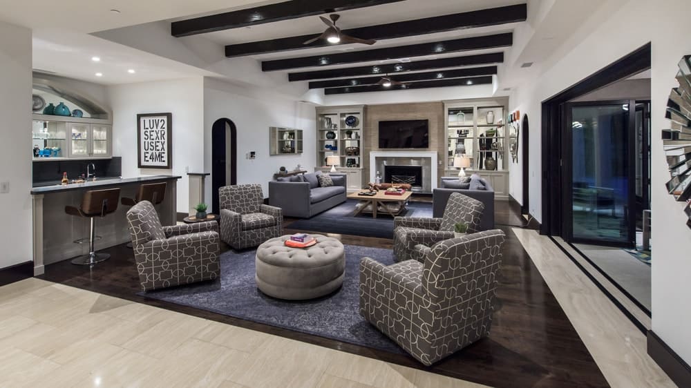 This is the spacious living room with sets of sofas and cushioned armchairs under a beamed ceiling. There is also a small wet bar on the side paired with a couple of stools. Image courtesy of Toptenrealestatedeals.com.