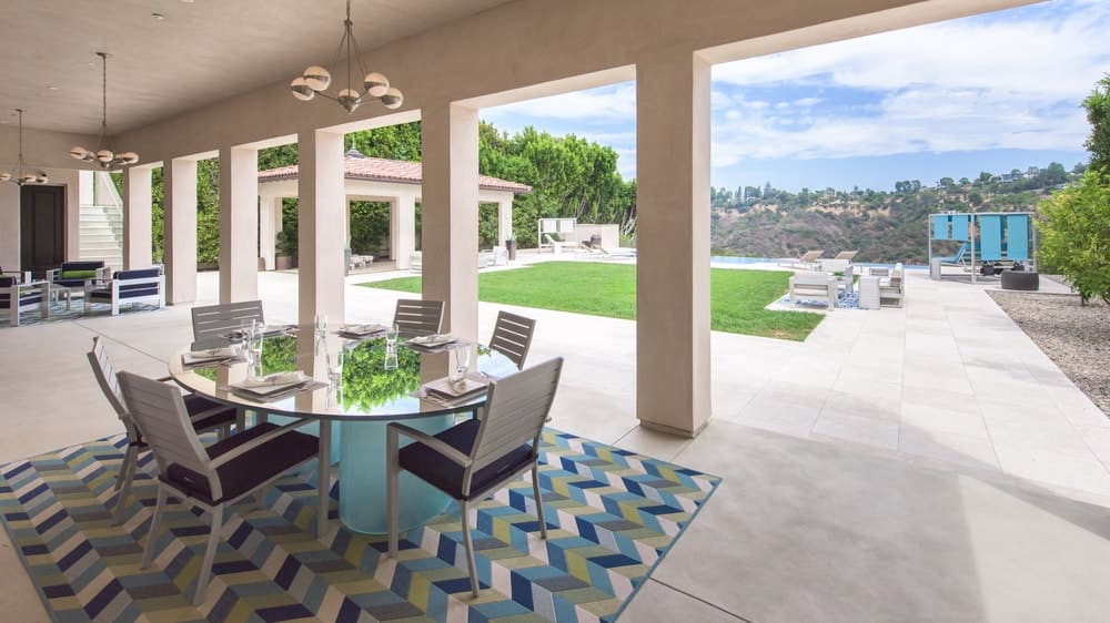 This is the covered patio at the side of pool area. This is supported by a row of pillars and open walls to brighten the glass-top outdoor dining set and outdoor kitchen. Image courtesy of Toptenrealestatedeals.com.