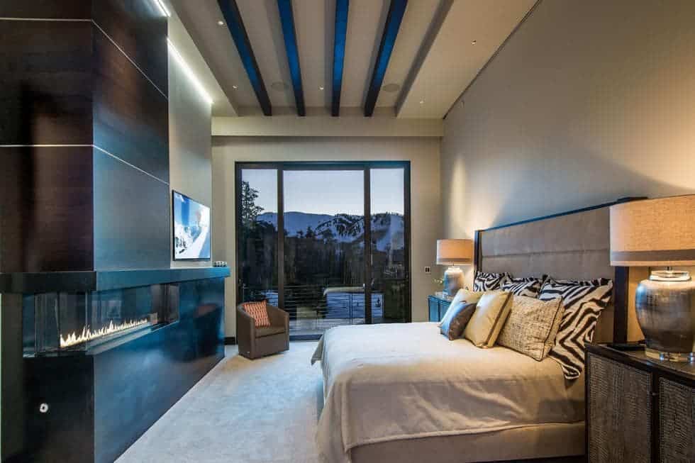 This is the bedroom with a tall beamed ceiling. The large bed has a large cushioned headboard across from the modern fireplace. Image courtesy of Toptenrealestatedeals.com.