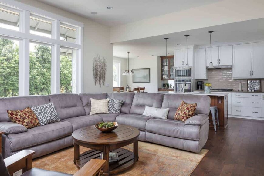 The living room has a gray V-shaped sectional, a round coffee table, and a three-panel window that invites natural light in.