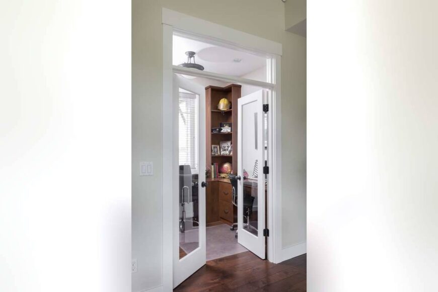 A white french door opens to the home office that comes with wooden built-ins and desks.