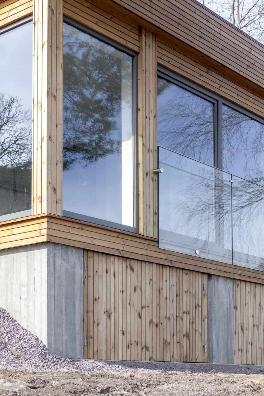 This is a close look at the exterior of the house with wooden shiplap elements, concrete elements and large glass walls and windows as well as railings.