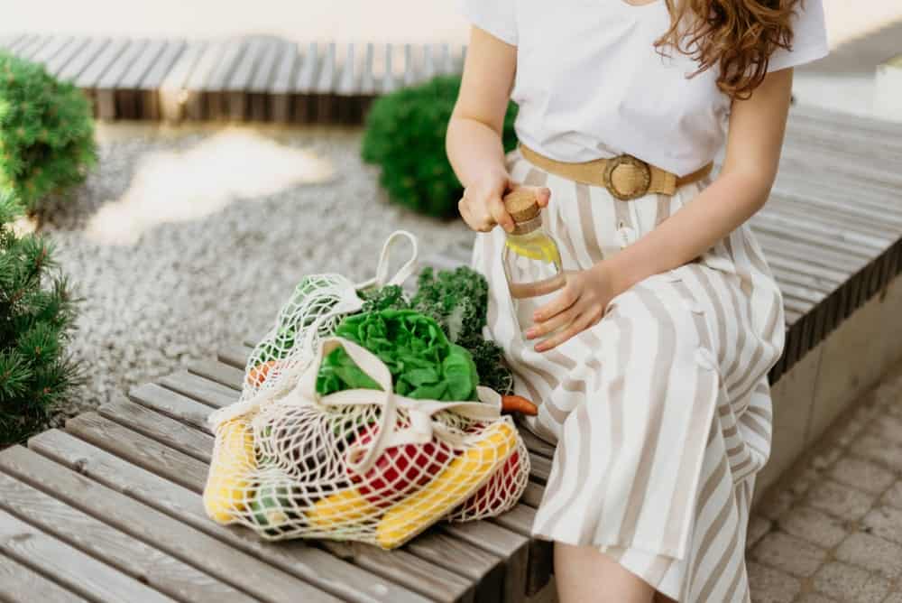 A woman sitting on a park bench with her groceries in an eco-friendly mesh bag.