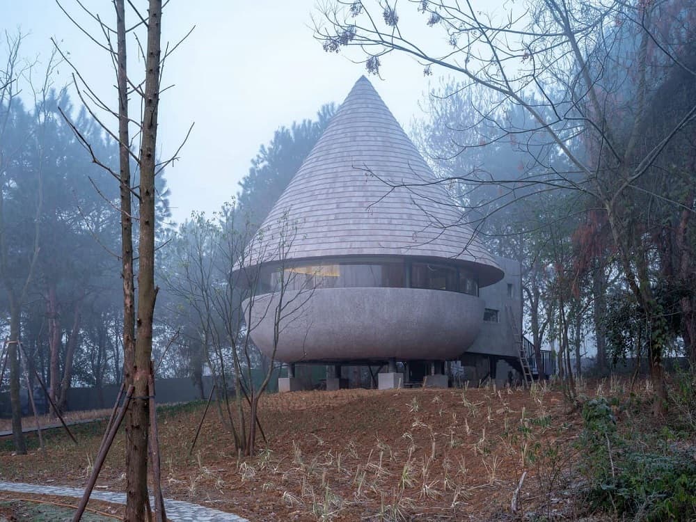 This is an exterior view of the house that has a unique design to its mushroom-like shape that has stilts underneath and a cone rooftop.