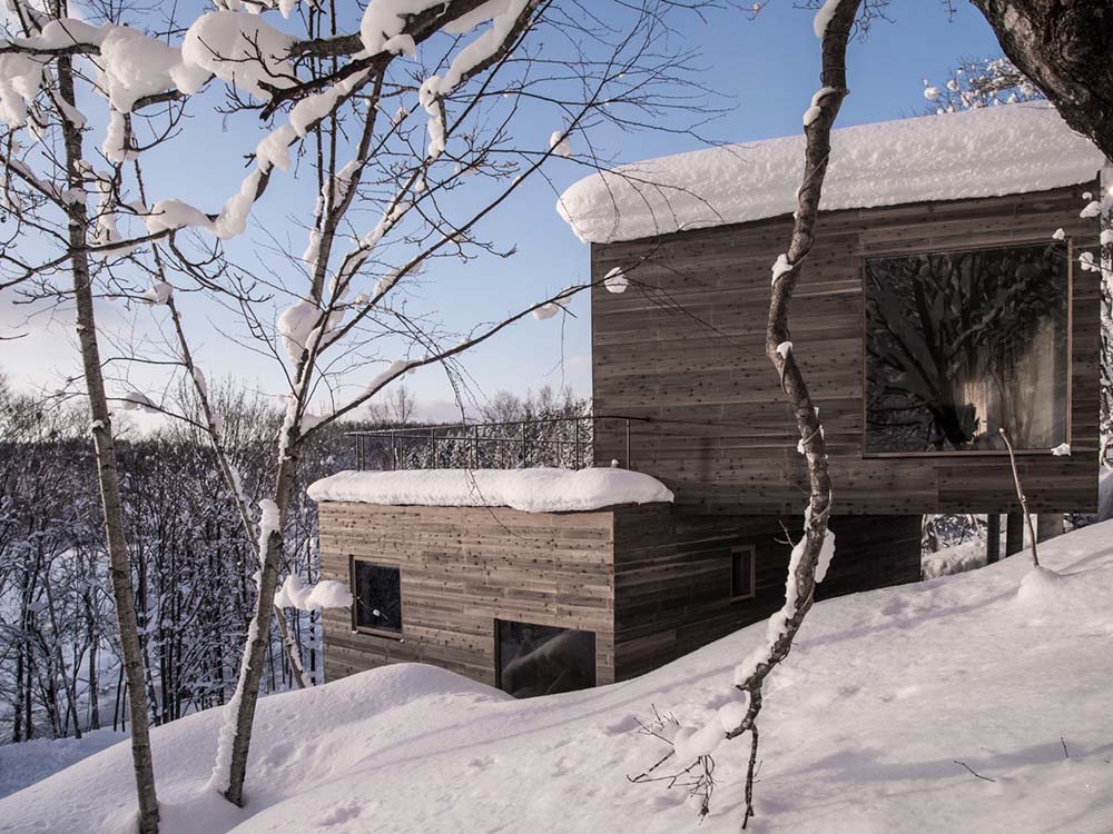 This is a view of the side of the house with dark brown textured exterior walls that stand out against the snowy landscape. You can also see here the balcony with thin railings.