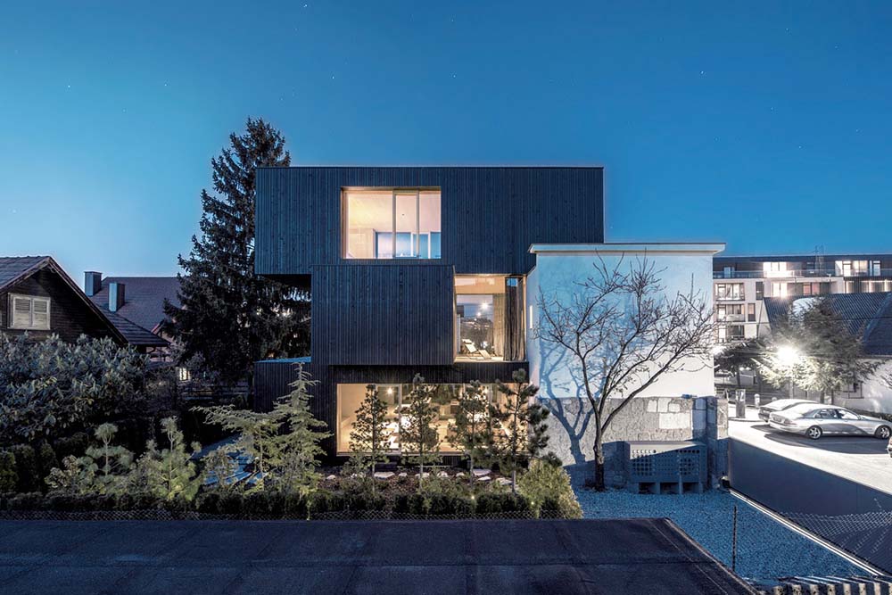 This is a view of the side of the house that showcases three box structures stacked on top of each other complemented by the glowing windows and landscaping of trees.