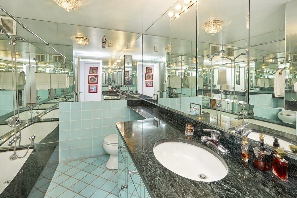 This bathroom has a dark marble counter to its vanity topped with a large wall mirror that extends to the toilet area on the far side. Image courtesy of Toptenrealestatedeals.com.