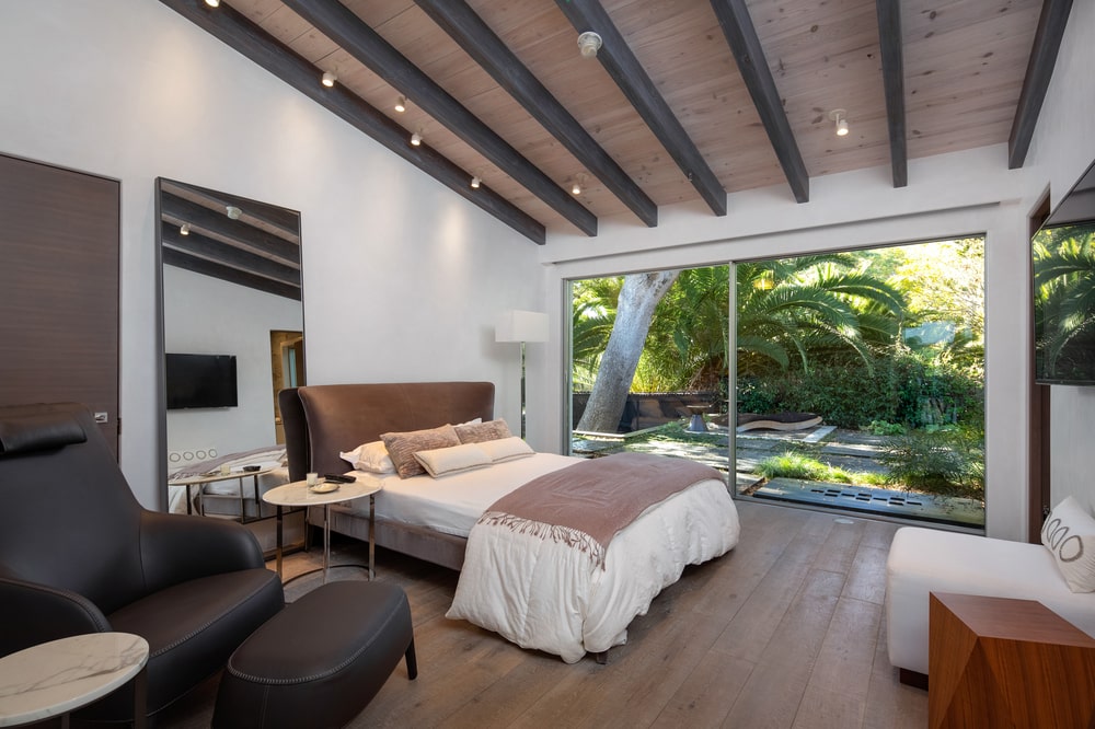 This bedroom has a bed with a large cushioned headboard by the large glass wall that shows the landscape outside. Image courtesy of Toptenrealestatedeals.com.