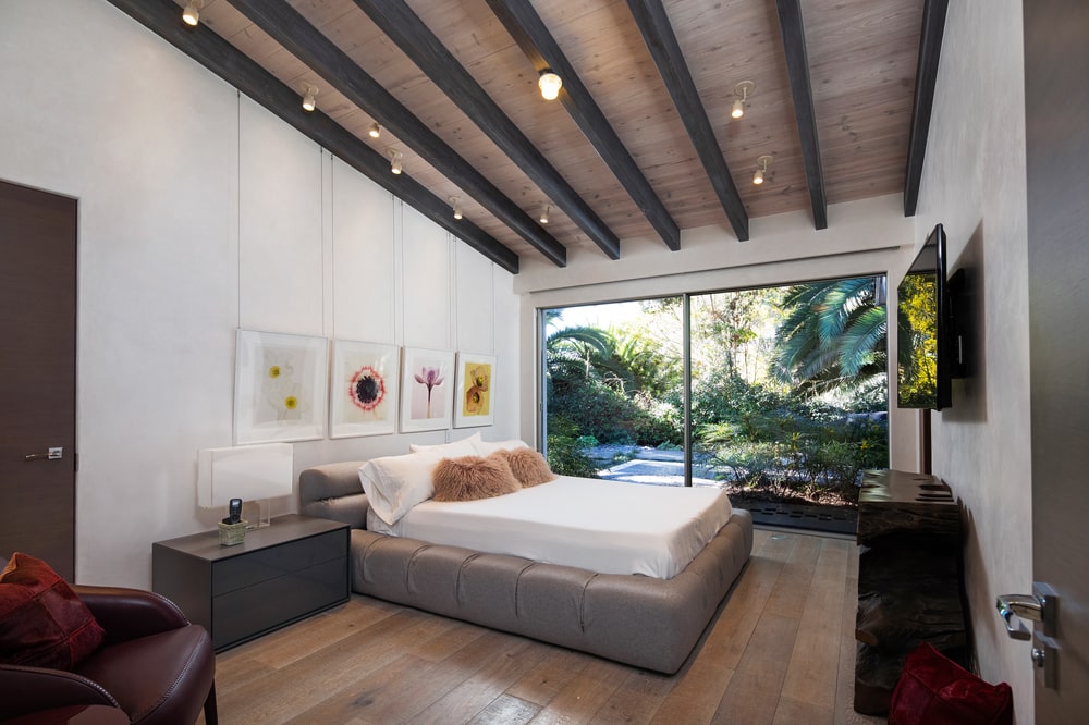 This is a bedroom that has a large tufted and cushioned bed frame for the bed topped with a shed beamed ceiling with decorative lighting. Image courtesy of Toptenrealestatedeals.com.